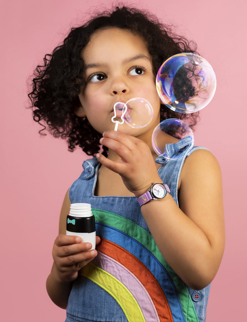 Joyful girl blowing bubbles while wearing a Tikkers analogue watch on a vibrant pink background